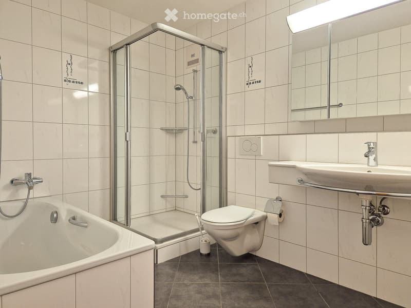 Modern bathroom with a bathtub, toilet, sink, mirror, and a glass-enclosed shower area. Features tiled walls and a dark tiled floor.