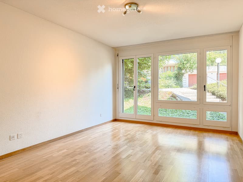Empty room with light wooden flooring, white walls, large windows, and a ceiling light fixture.