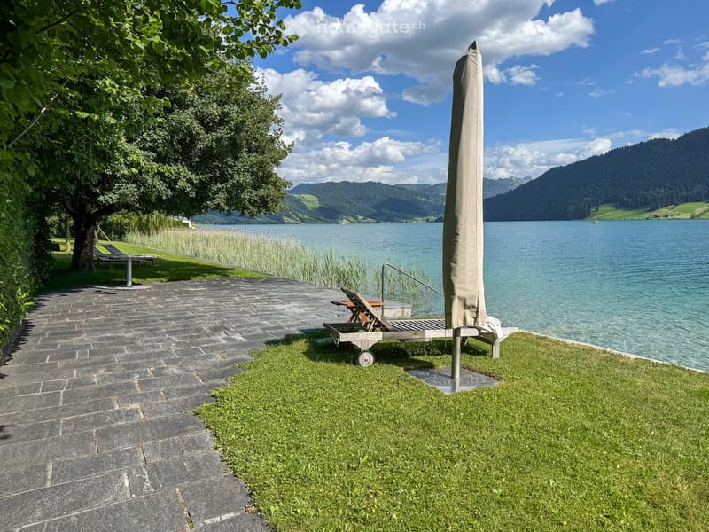 Outdoor area with a stone-paved pathway, a grassy lawn, a large umbrella, a wooden chair, and a table near a lake with mountains in the background.