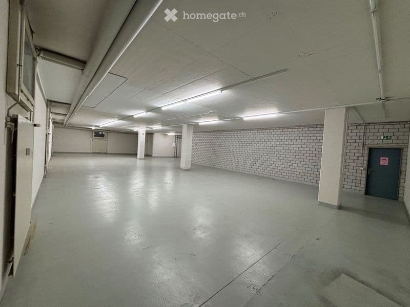 Empty, spacious room with concrete floors and white brick walls, illuminated by overhead fluorescent lighting. The room features several white support columns and a single grey door on the right side.