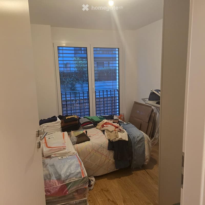 Small bedroom with a single bed, covered with a patterned quilt and various folded clothes. An ironing board and iron are positioned near the bed, along with several storage bins and a stack of folded towels.
