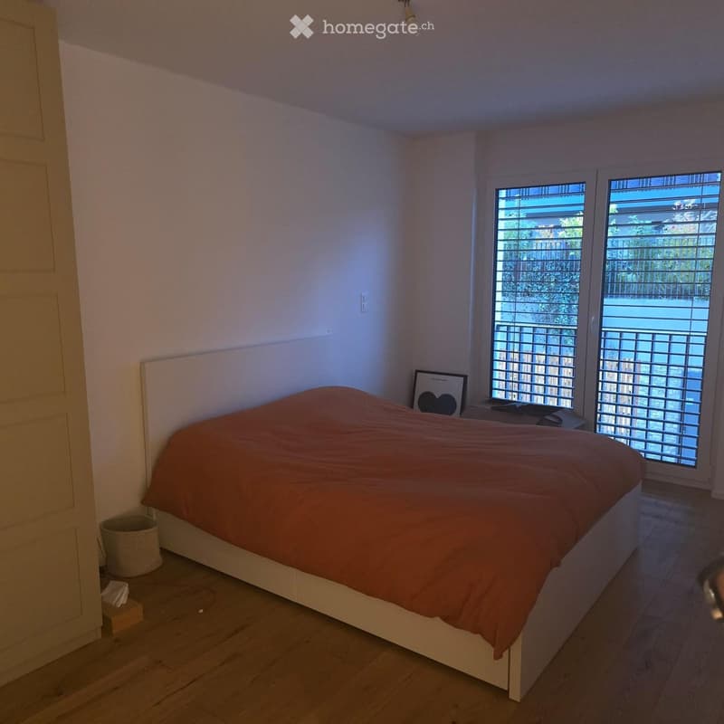 Cozy bedroom with a wooden bed, orange bedspread, nightstand, and a view through large windows with blinds.