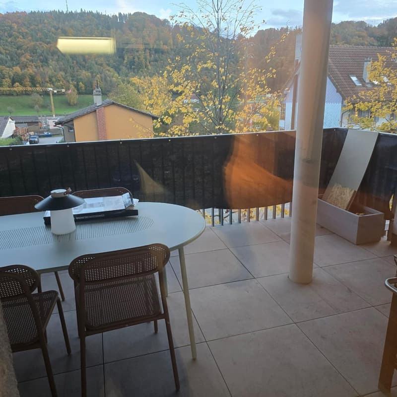 Balcony with a round table, two chairs, and a lamp, overlooking a scenic landscape with trees and houses.