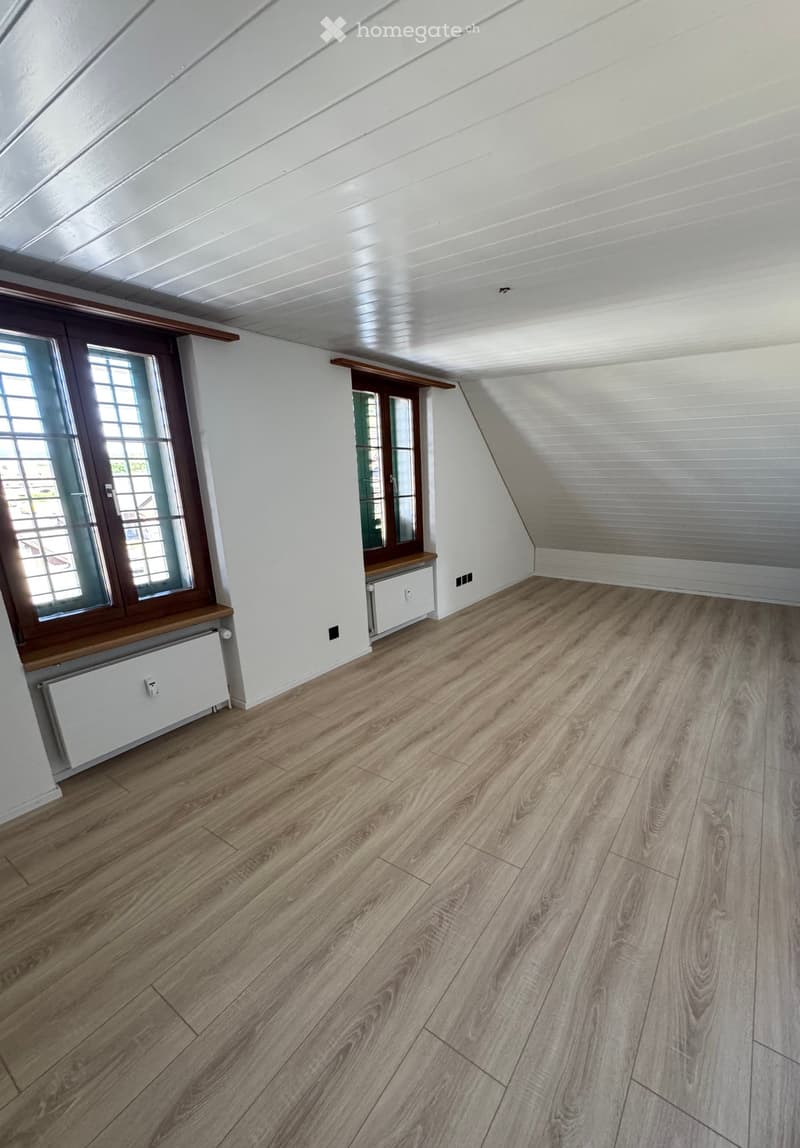 Empty attic room with wooden flooring, white walls, and large windows with wooden frames. The ceiling has exposed wooden beams and a slanted design.