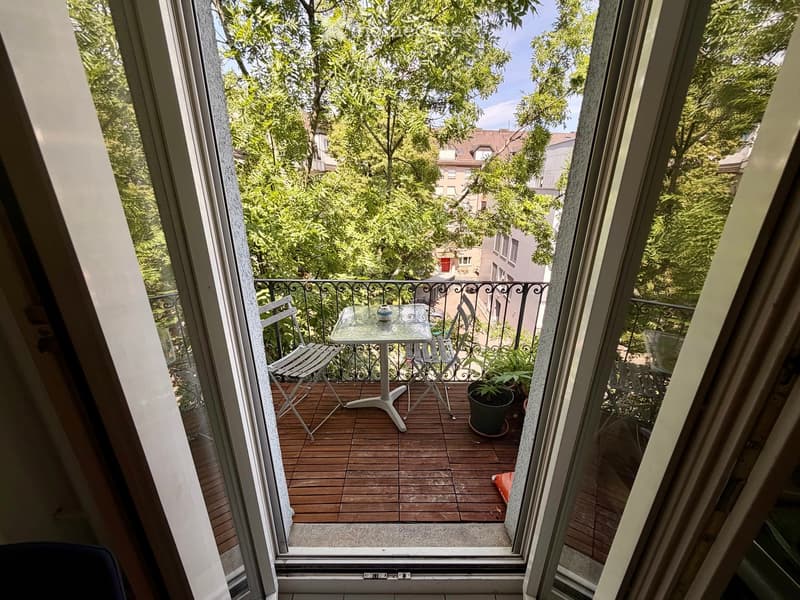 Balcony with wooden decking, white table, two chairs, and a potted plant, overlooking a leafy urban area.