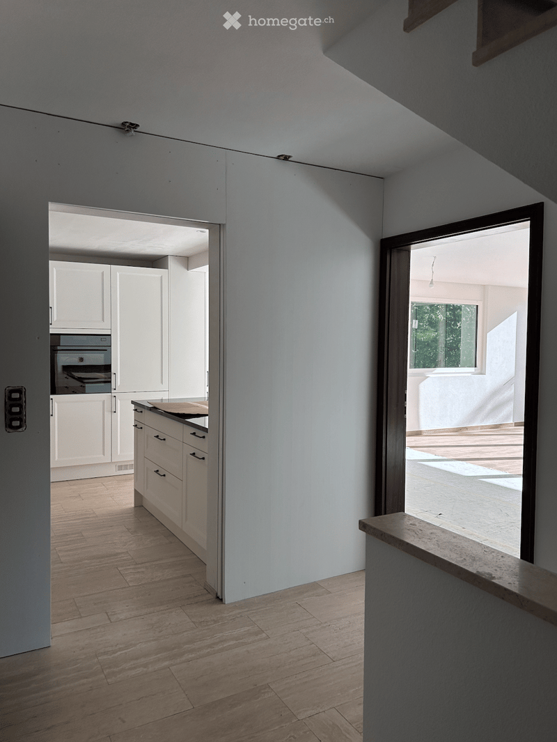 Bright, minimalist hallway with light wooden flooring, white cabinets, and an open doorway leading to a kitchen area with an oven visible.
