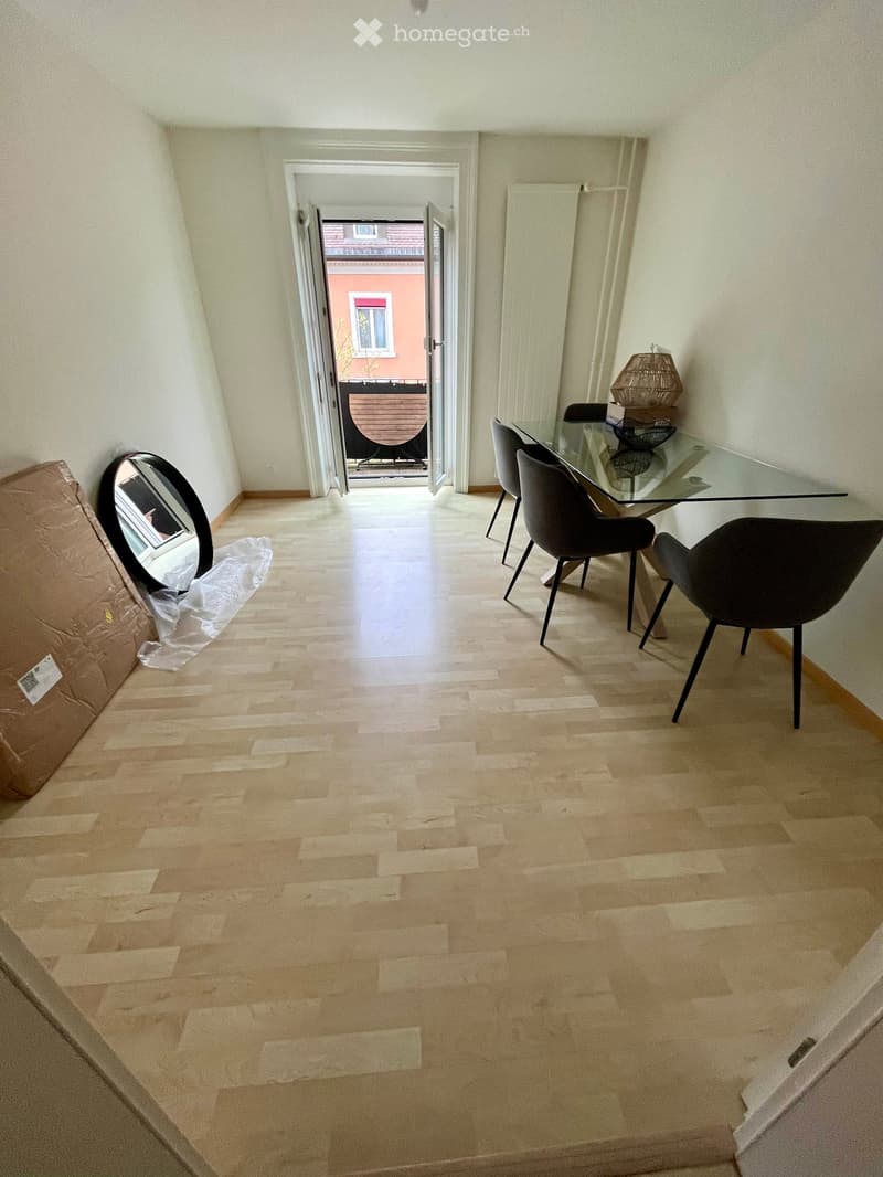 Dining area with a glass table, four modern chairs, a wicker lamp, and a large round mirror leaning against the wall. The room has wooden flooring and leads to a balcony.