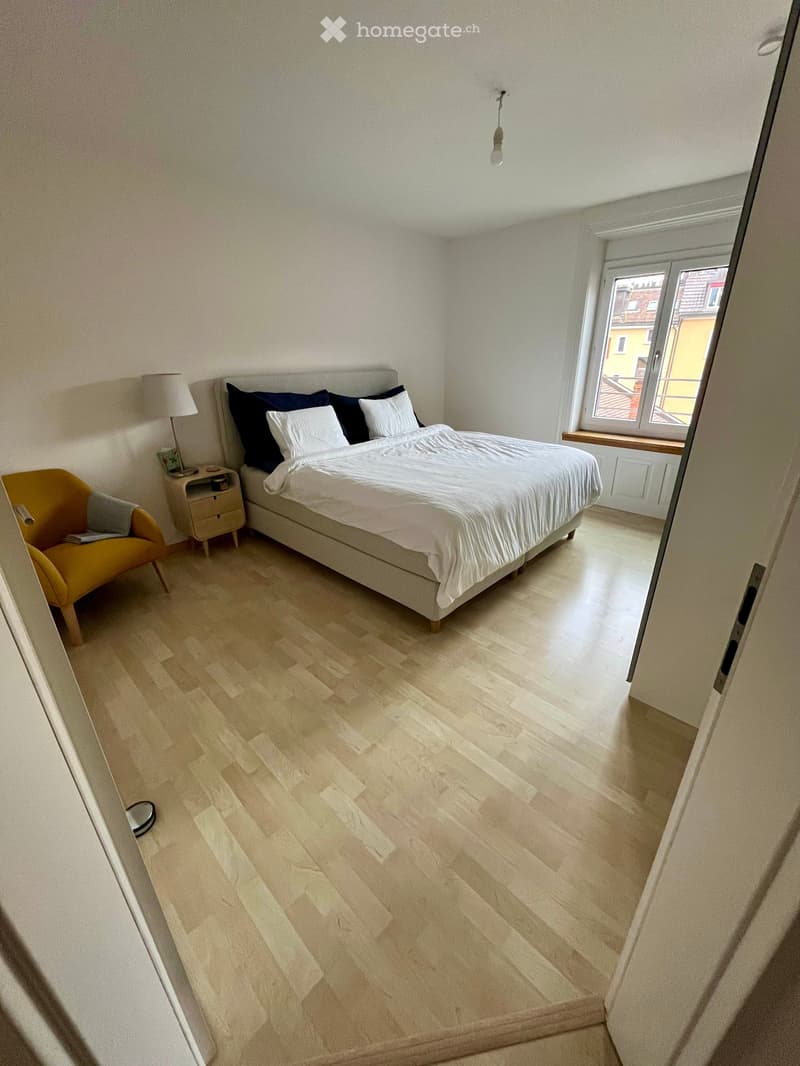 Bright bedroom with wooden flooring, a large bed with white and dark blue bedding, a nightstand with a lamp, and a yellow armchair.
