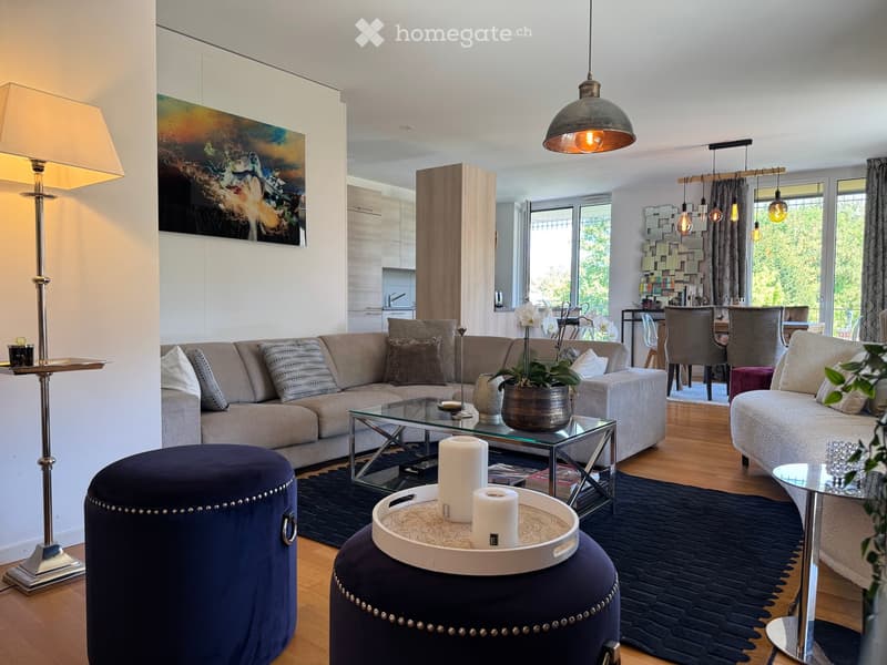 Modern living room with a grey sectional sofa, glass coffee table, blue velvet stools, pendant lights, floor lamp, and a dining area with chairs and a table.