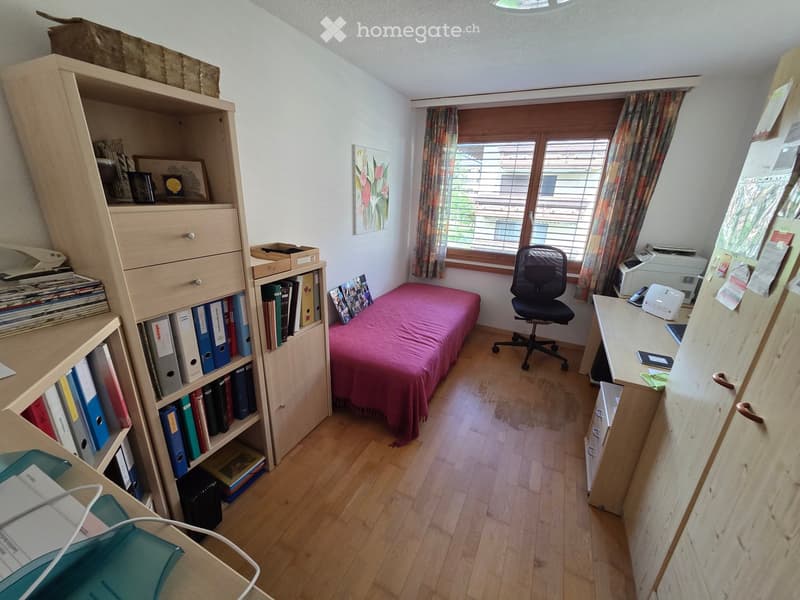 A small, furnished room with a bed, desk, chair, bookshelf, and window with curtains.
