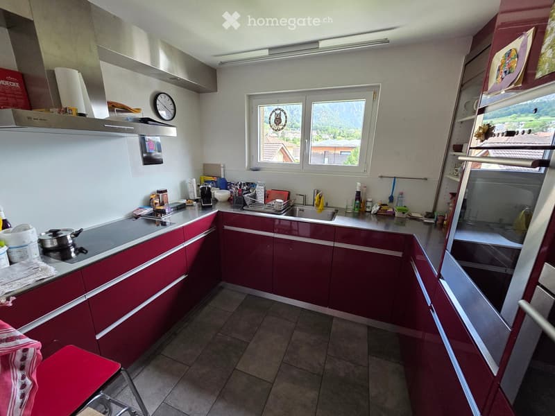 Modern kitchen with red cabinets, white countertops, and stainless steel appliances. Features a large window with a scenic view, a sink, stove, and various kitchen items on the counter.