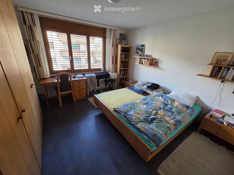 Cozy bedroom with a single bed, desk, chair, bookshelf, and nightstand. The room features wooden furniture and large windows with curtains.