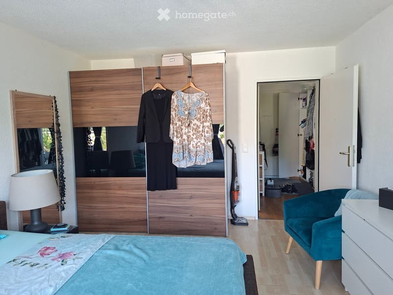 A well-furnished bedroom with a large bed, a wooden wardrobe, a teal armchair, a lamp, and a mirror. The room features wooden flooring and a partially visible adjacent space.