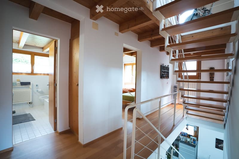 Bright hallway with wooden floors and exposed wooden beams. Features a staircase leading to an upper level, with visible rooms including a bathroom and bedroom.