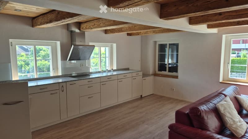 Open-plan kitchen and living area with light-colored cabinets, granite countertop, red leather sofa, and exposed wooden beams on the ceiling.