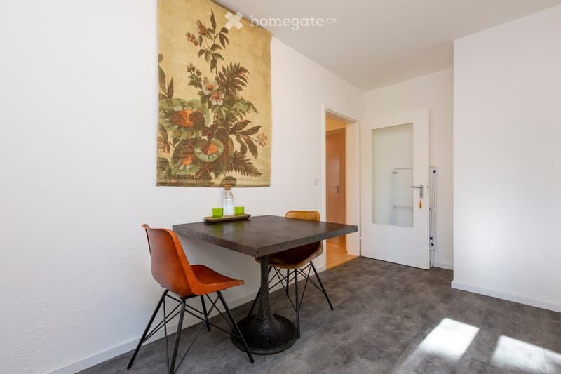 A modern, minimalist room with a wooden table, two orange chairs, a bottle, and cups on the table. A large floral tapestry hangs on the wall, and a doorway leads to another room.