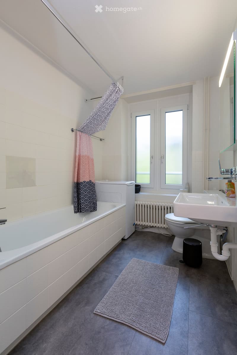Modern bathroom with a bathtub, toilet, sink, mirror, radiator, and a grey bathroom rug. The room features white tiled walls and a grey tiled floor, with a window providing natural light.