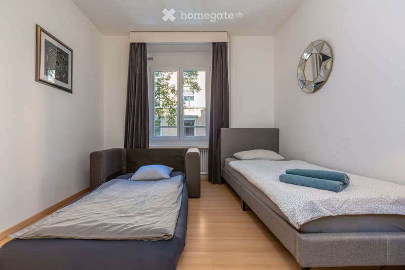 Cozy bedroom with two single beds, a mirror, and a picture frame on the wall. The room features wooden flooring and large windows with curtains.