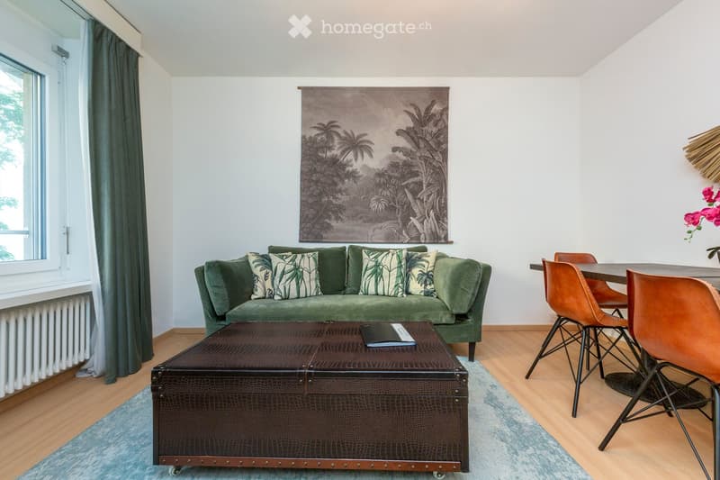 Living room with green velvet sofa, coffee table, dining table with chairs, large painting, and a flower vase on the side table.