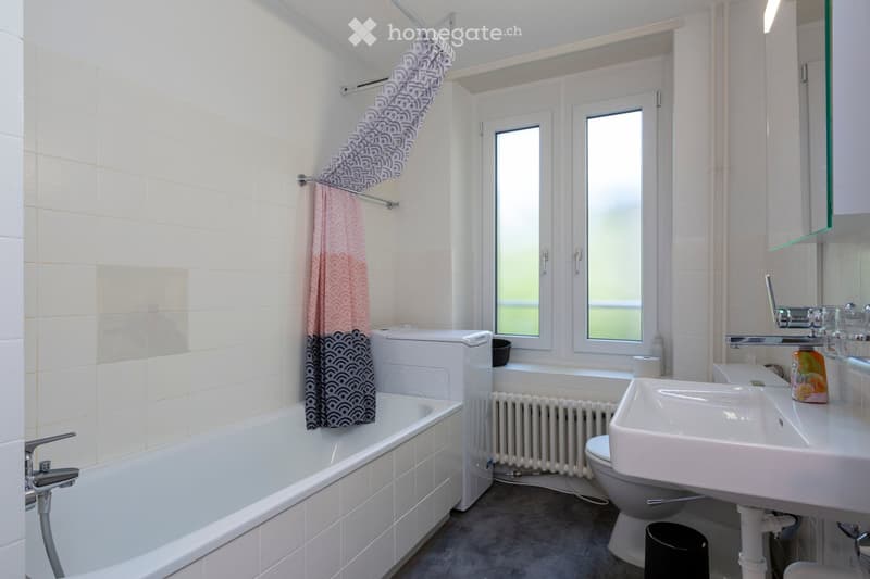 Modern bathroom with white tiled walls, a bathtub, a sink, a toilet, a radiator, and a shower curtain. The room features a window with frosted glass panes and a small shelf with toiletries.