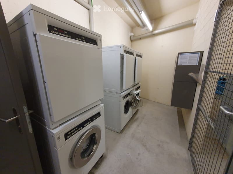A small, well-lit room with multiple washing machines and dryers stacked together, a screen mounted on the wall, and a metal cage on the right side.