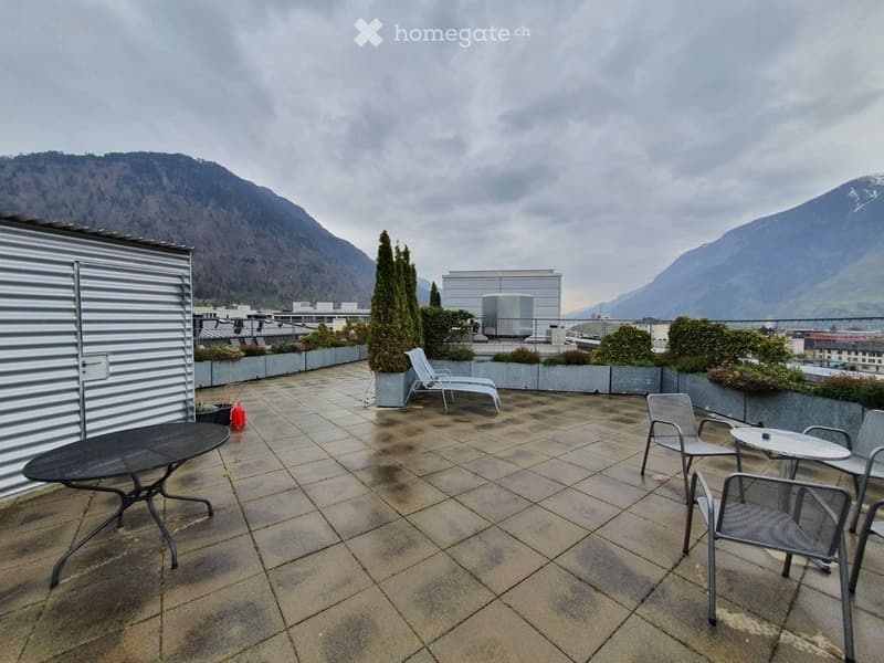 Roof terrace with tiled floor, lounge chairs, tables, and potted plants, overlooking a cityscape with mountains in the background.