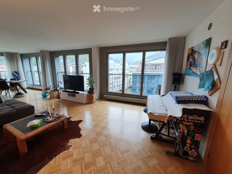 A modern, well-lit living room with wooden flooring, large windows, and a balcony view. The room features a sofa, coffee table, television, and a guitar on a stand.