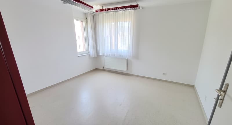 Empty room with white walls, light-colored floor, window with white curtains, radiator, and a door with a handle.