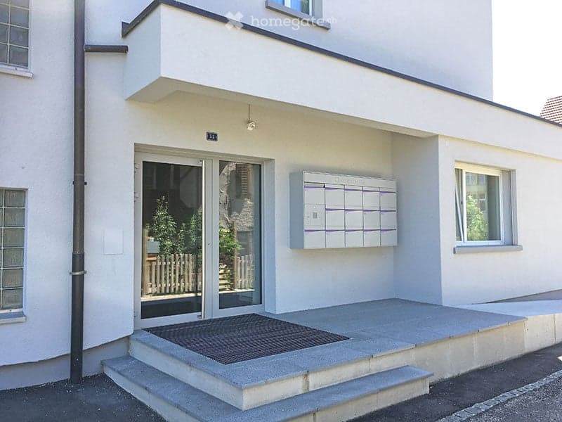 Modern two-story building with a raised ground floor, white facade, and a small porch with glass doors leading to an interior space.