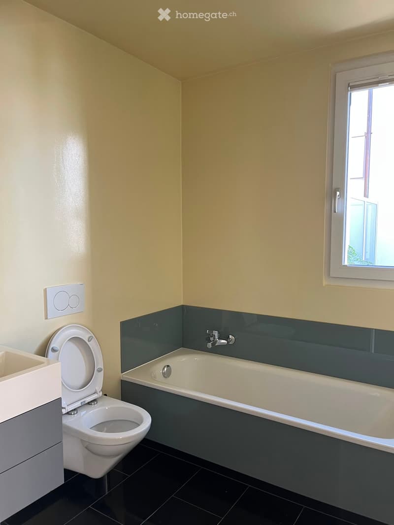Bathroom with a toilet, bathtub, and sink. Light yellow walls with a dark green accent border near the bathtub. Black tiled floor and a window providing natural light.