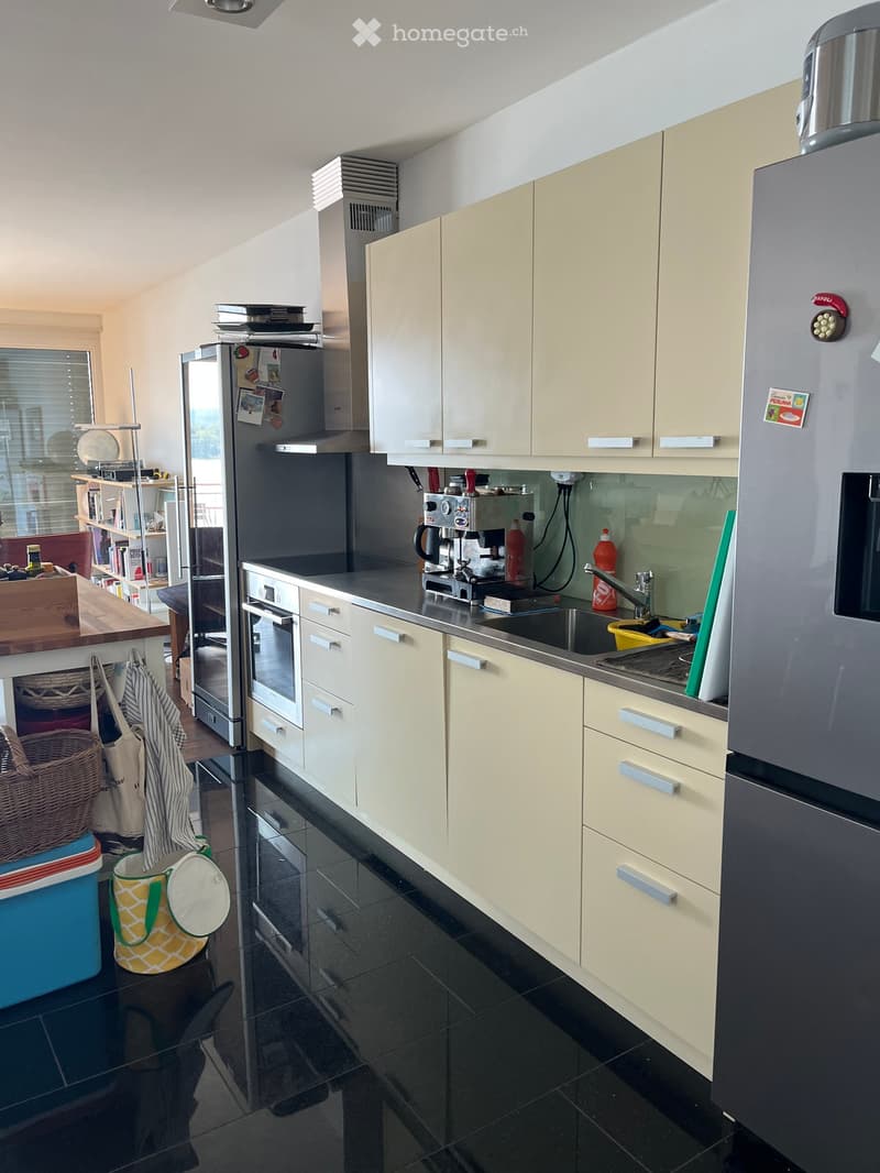 Modern kitchen with light-colored cabinets, stainless steel appliances, and black tiled flooring. Features include a refrigerator, oven, sink, coffee maker, and various storage items.