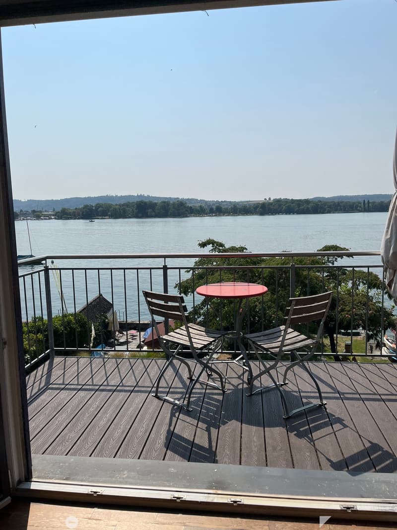 Balcony with wooden decking, metal railing, and a red round table with two wooden chairs, overlooking a lake.