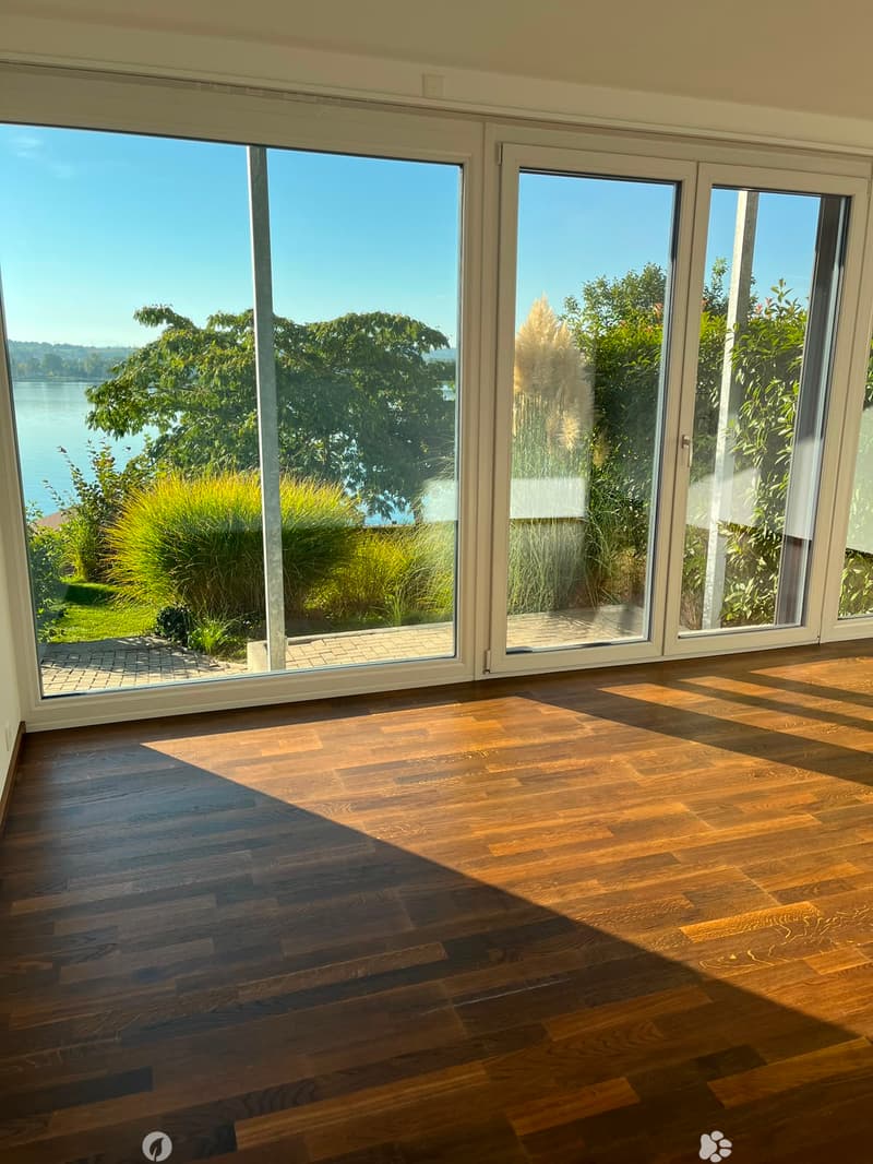 Empty room with wooden flooring and large sliding glass doors leading to a view of a lake and garden.