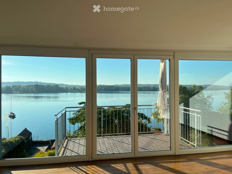 Large sliding glass doors open to a wooden deck with a metal railing, overlooking a serene lake with trees and a small boat visible.