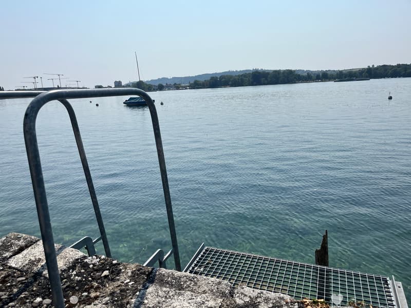 View of a calm lake with a small boat, distant construction cranes, and a distant shoreline with trees and buildings.
