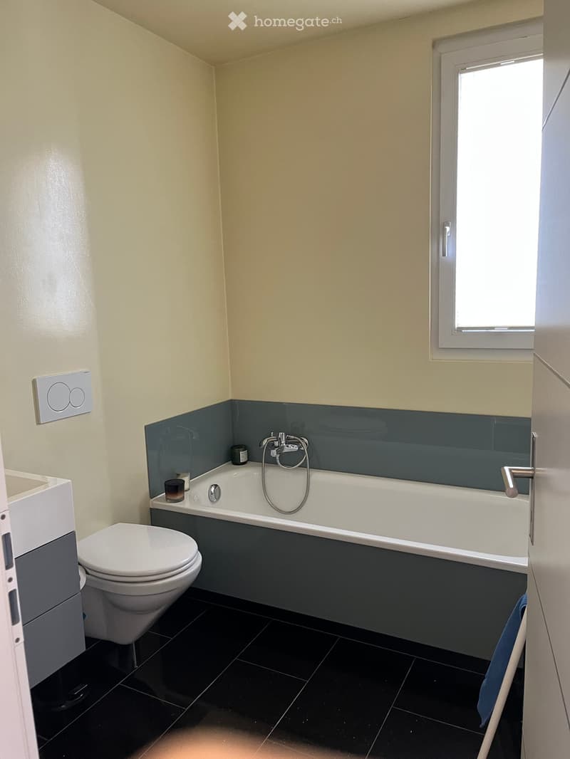 Modern bathroom with a bathtub, toilet, sink, and a small window. The walls are painted in a light beige color with a dark blue accent around the bathtub area. The floor is covered with black tiles.