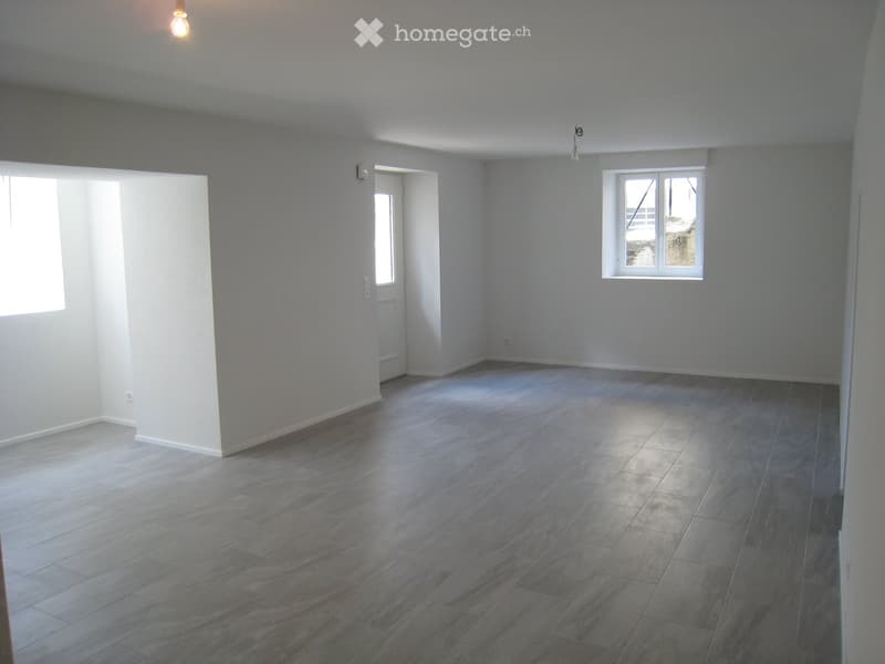 Empty room with light grey walls, wooden flooring, and two windows allowing natural light. No furniture or equipment is present.