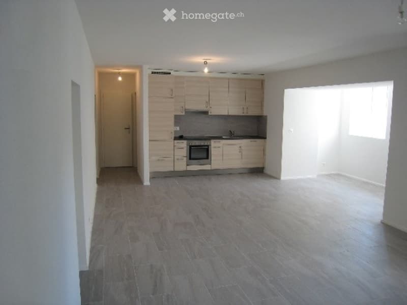 Modern kitchen with light wood cabinets, stainless steel oven, and a large window letting in natural light. The floor is tiled in a dark grey color.
