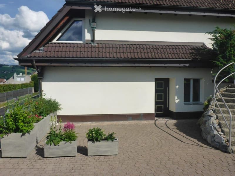 Single-story house with white walls, dark brown roof, and a front entrance. The house features a small garden area with raised planters and a curved brick pathway leading to the entrance.