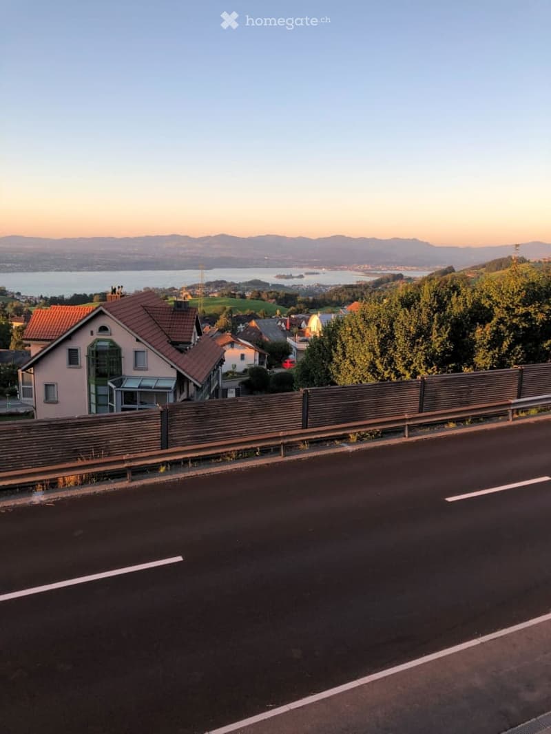 A road runs alongside a wooden fence, with houses and a scenic view of a lake and distant mountains in the background.