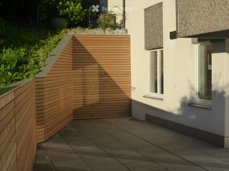 Wooden fence and tiled floor on a raised terrace, adjacent to a house with windows and a small garden.