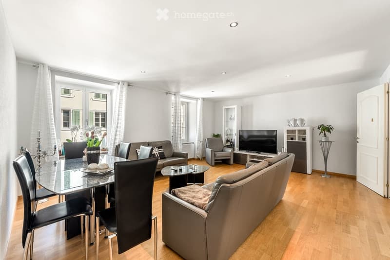 Modern living and dining area with light wooden flooring, large windows with white curtains, a grey sofa, a glass dining table with black chairs, a television, and a small plant on a side table.
