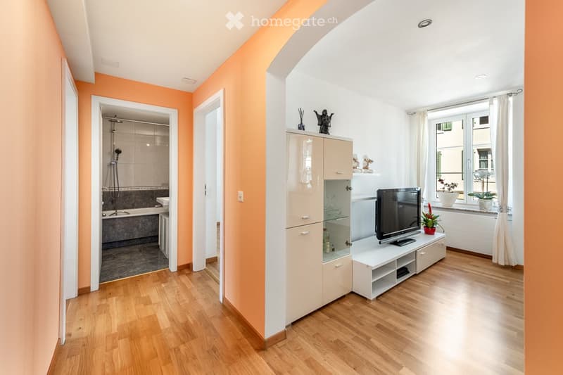 A modern living room with wooden flooring, white walls, and a large window with white curtains. A flat-screen TV is placed on a white cabinet, accompanied by decorative items and a small plant on the windowsill. An open doorway leads to a bathroom with a