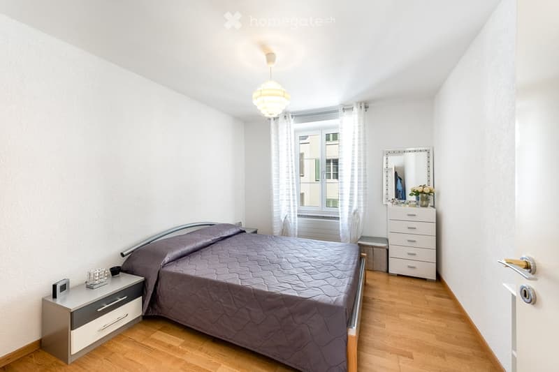 Cozy bedroom with wooden flooring, a neatly made bed with dark bedding, a nightstand with a lamp, a dresser with a mirror, and a small flower vase on top.