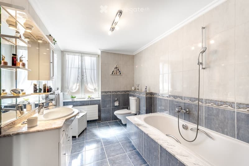 Elegant bathroom with a double sink, bathtub, toilet, and a large mirror. The space features modern fixtures, tiled walls, and a window with curtains.