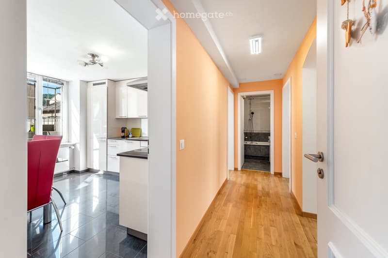 Bright hallway with wooden flooring, leading to a kitchen and bathroom. The kitchen features white cabinets, a countertop, and a red chair. The bathroom has a bathtub and a shower area.