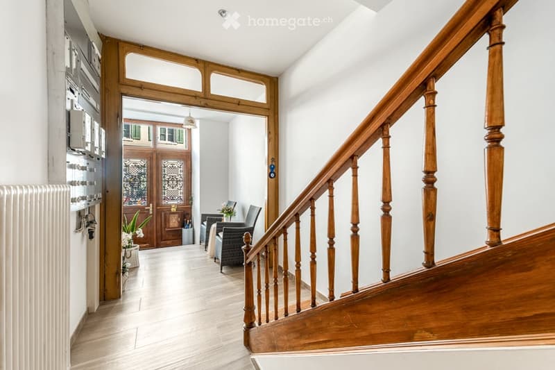 Spacious hallway with wooden staircase, white walls, and a view into a living area with seating and a door leading outside.