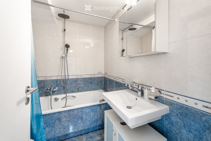 Modern bathroom with a bathtub, sink, mirror, and shower head. Blue tiles line the lower half of the walls, with white tiles above. A cabinet is positioned under the sink.