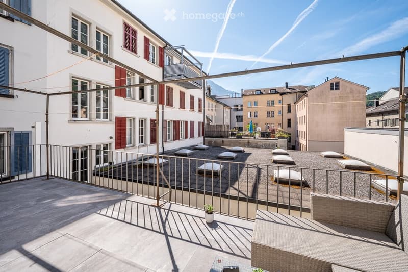Rooftop terrace with modern furniture, overlooking a cityscape with traditional buildings and distant mountains.