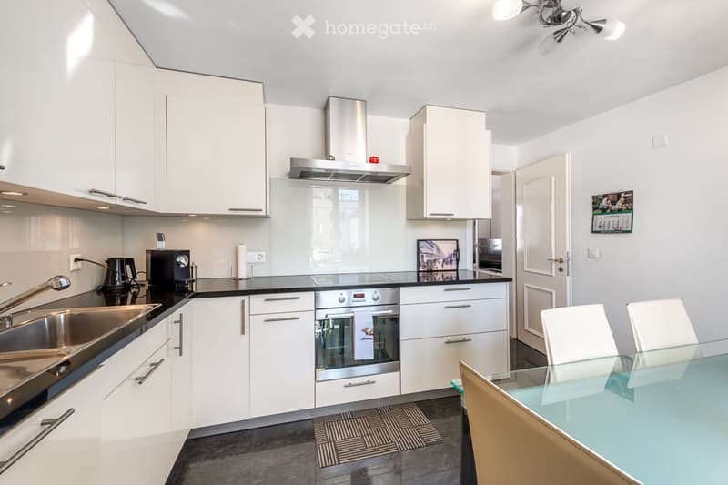 Modern kitchen with white cabinets, stainless steel appliances, and a black countertop. Includes a sink, coffee maker, oven, range hood, and a small dining area with a glass table and white chairs.